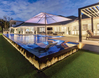 Luxury backyard with illuminated pool, pink umbrella, loungers, and modern house with pergola at dusk