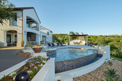 Modern luxury home with curved swimming pool, white architecture, and manicured landscaping overlooking green hillside
