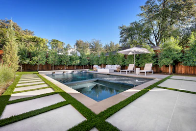 Modern luxury backyard with rectangular swimming pool, spa, lounge seating, and manicured landscaping surrounded by wooden fence and mature trees