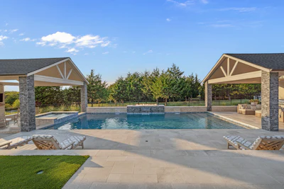 Luxury backyard with resort-style swimming pool, dual stone pavilions, lounge chairs, and manicured lawn under blue sky
