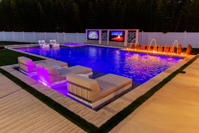 Luxury backyard pool at night with blue water, glowing lounge chairs, fountain features, and illuminated display screens