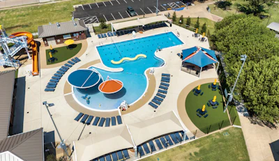 Aerial view of a community aquatic center with Olympic-sized pool, splash pad, water slides, and recreational facilities surrounded by landscaping and parking.