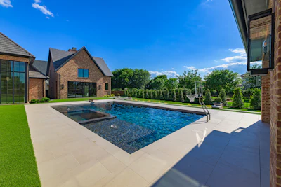 Luxury modern brick home with rectangular swimming pool, manicured lawn, and hedged landscaping under clear blue sky