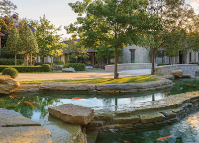 Serene courtyard with stone buildings, mature trees, manicured lawn, and koi pond with stepping stones