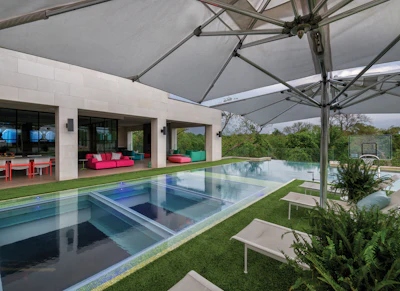 Modern luxury home with infinity pool, white architecture, pink seating, and retractable shade structure overlooking landscaped grounds
