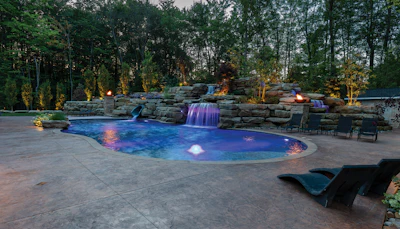 Luxurious backyard pool with illuminated waterfall, rock features, and lounge seating surrounded by trees at dusk