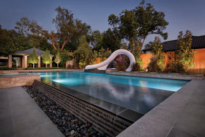 Modern luxury backyard pool with curved white slide, blue LED lighting, and manicured landscape at twilight