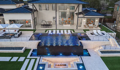 Aerial view of modern luxury home with blue-tiled pool, spa, fire pit, and manicured landscaping