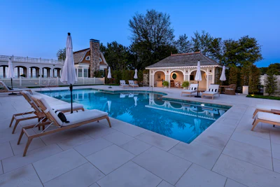 Luxury poolside estate with blue water, white lounge chairs, cream-colored buildings, and manicured landscaping at dusk