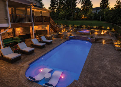 Illuminated backyard with rectangular swimming pool, lounge chairs, and multi-level deck at dusk