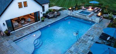 Aerial view of luxury backyard with rectangular swimming pool, spa, patio seating, and modern white house with blue roof