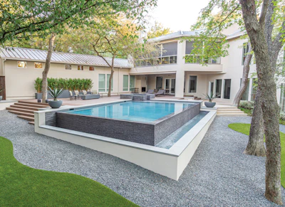 Modern luxury home with rectangular swimming pool, gray stone patio, manicured lawn, and mature trees