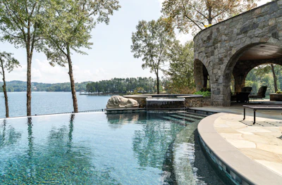 Luxury waterfront infinity pool with stone bridge overlooking a serene lake surrounded by trees