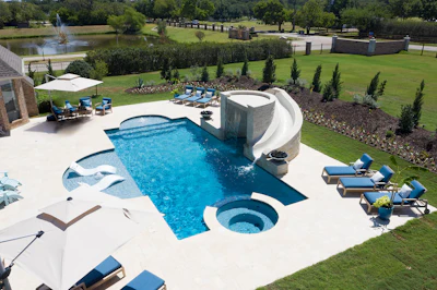 Luxury backyard pool complex with curved waterslide, spa, lounge seating, and manicured grounds overlooking a pond.