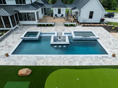 Aerial view of luxury home with rectangular swimming pool, spa, and stone patio surrounded by manicured lawn and modern architecture.
