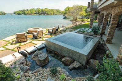 Luxury lakefront home with infinity pool, stone patio, fire pit, and waterfront seating overlooking calm blue water and tree-lined shore.