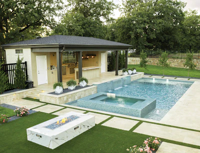 Modern luxury backyard with infinity pool, spa, fire pit, and contemporary pool house surrounded by manicured lawn and mature trees.