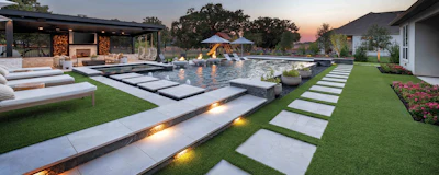 Luxury backyard with illuminated pool, lounge seating, fire pit, and manicured lawn at dusk