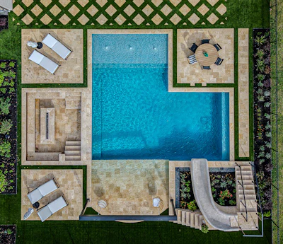 Aerial view of a luxury residential pool complex with blue swimming pool, stone patio, lounge chairs, and manicured green landscaping