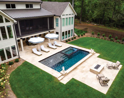 Aerial view of modern home with rectangular pool, patio lounge area, and manicured lawn surrounded by trees
