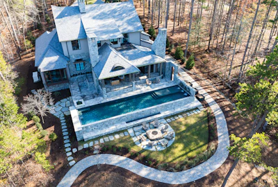 Aerial view of luxury stone mansion with heated pool, curved driveway, and fire pit surrounded by pine forest