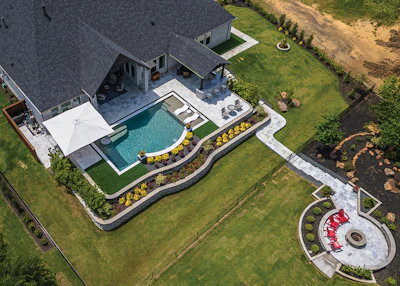 Aerial view of luxury home with curved swimming pool, patio, manicured lawn, and circular fire pit area