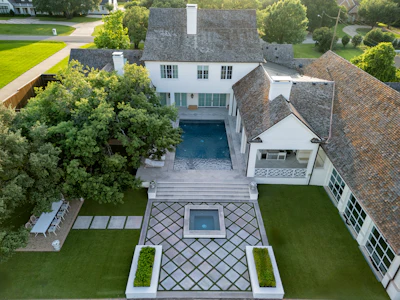 Aerial view of modern white house with gray roof, circular driveway, courtyard with geometric patterned pavers, and manicured lawn surrounded by trees.