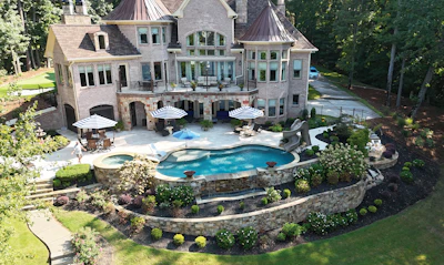 Aerial view of luxury mansion with curved swimming pool, spa, stone patio, white umbrellas, and manicured landscaping surrounded by trees