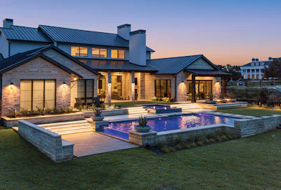 Modern luxury home with metal roof, stone exterior, illuminated windows, and lit swimming pool at twilight