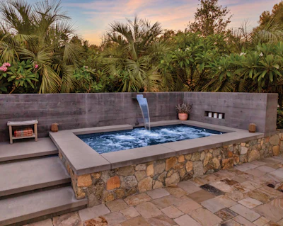 Luxury sunken hot tub with stone base and waterfall feature surrounded by tropical landscaping at sunset
