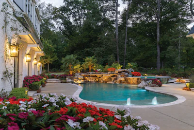 Luxury backyard with illuminated pool, stone waterfall, colorful flower gardens, and white house at dusk