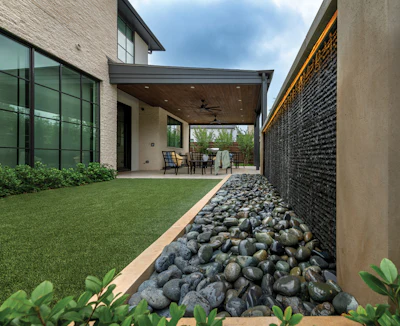 Modern home exterior with covered patio, manicured lawn, decorative rock border, and contemporary architecture featuring large windows.