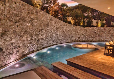 Luxury backyard with illuminated swimming pool, spa, wooden deck, and stone accent wall at dusk