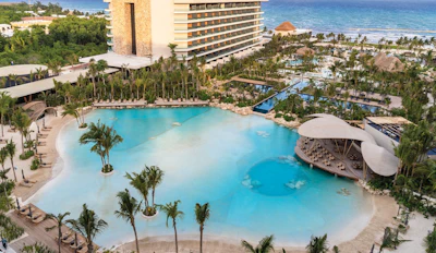 Aerial view of luxury beachfront resort with turquoise lagoon pool, palm trees, high-rise hotel buildings, and ocean coastline