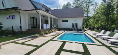 Modern white farmhouse with rectangular pool, geometric patio design, and lounge chairs overlooking manicured lawn and trees.