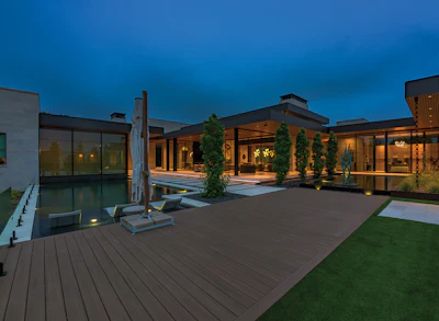 Modern luxury home courtyard at dusk with illuminated building, reflecting pool, manicured lawn, and landscaping.