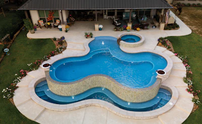Aerial view of luxury backyard with curved swimming pool, spa, and patio near modern home with metal roof.