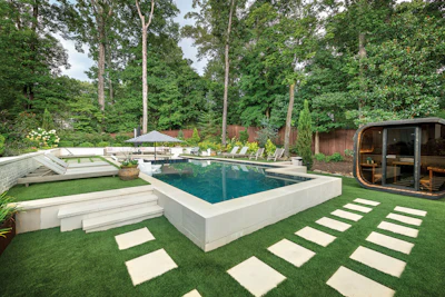Modern luxury backyard with rectangular pool, white pavers, manicured lawn, lounge chairs, umbrella, sauna, and mature trees.