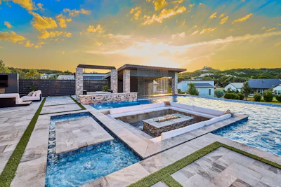 Luxury modern home with infinity pool, fire pit, and architectural stonework at sunset overlooking hillside landscape