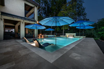 Luxury backyard with illuminated swimming pool, blue umbrellas, lounge chairs, and modern house at dusk.