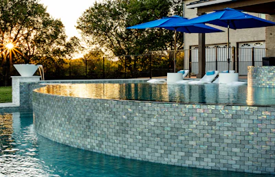 Modern luxury backyard pool with turquoise water, mosaic tile walls, blue umbrellas, and sunset lighting.