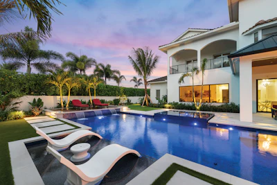 Luxury backyard with illuminated pool, modern white house, palm trees, and lounge chairs at dusk