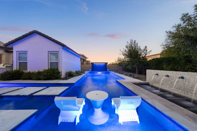 Modern home with illuminated blue swimming pool at dusk, featuring white lounge chairs and contemporary architecture