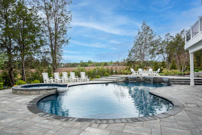 Luxury backyard with curved swimming pool, hot tub, white Adirondack chairs, and manicured landscaping overlooking open fields
