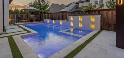 Modern backyard with illuminated rectangular swimming pool, concrete patio, manicured lawn, wooden fence, and mountain views at twilight.