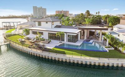 Modern waterfront home with infinity pool, manicured lawn, and palm trees overlooking calm water