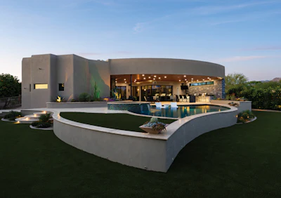 Modern curved concrete home with illuminated interior, curved swimming pool, and landscaped lawn at dusk