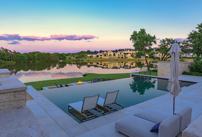 Luxury waterfront estate with infinity pool, lounge seating, and manicured lawn overlooking river at sunset