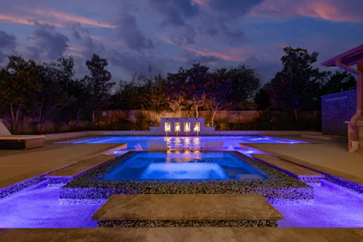 Luxury pool area with blue LED lighting and illuminated structure at dusk, surrounded by landscaping and trees