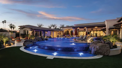 Luxury desert home with illuminated resort-style pool, multiple buildings, and manicured landscaping at twilight